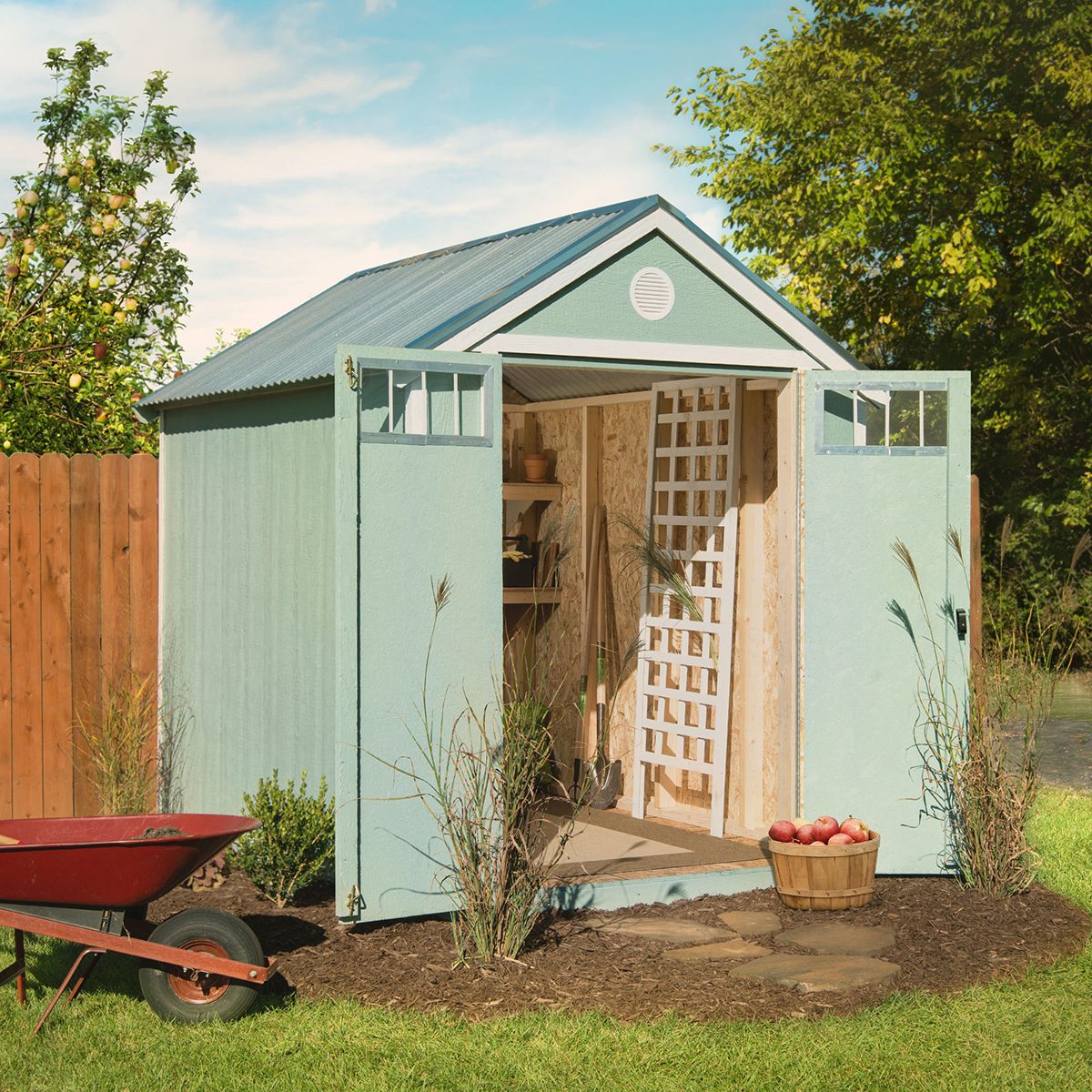 Rustic green garden shed with open double doors, metal roof, and interior storage, surrounded by plants and gardening tools.